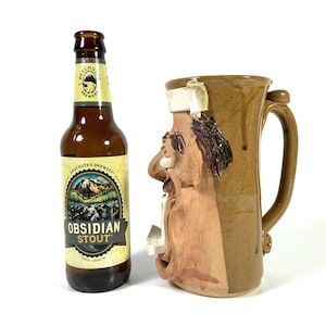 May include: A brown glass bottle of Deschutes Brewery Obsidian Stout next to a brown ceramic mug with a face design. The mug has a handle and a unique, textured appearance. The bottle has a yellow label.