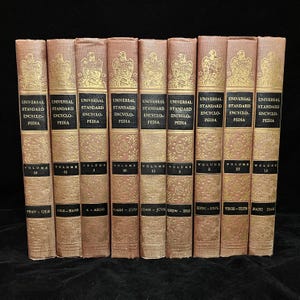 May include: A collection of vintage encyclopedias with tan covers and gold lettering. Each book displays the title "Universal Standard Encyclopedia" and volume numbers. The spines feature an eagle emblem. The volumes are arranged in a row.