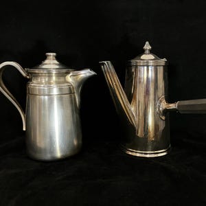 May include: Two silver-toned metal coffee pots with polished finishes. The left pot features a curved handle and a lid with a small knob. The right pot has a tall, cylindrical body, a long spout, a flat lid with a finial, and a straight dark handle.
