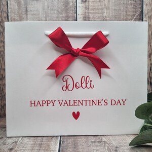 May include: White gift bag with a red bow and the text "Dolli Happy Valentine's Day" with a red heart.