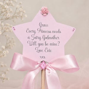 Op de afbeelding: Een stervormige roze toverstaf met een kleine roos aan de bovenkant, twee edelstenen aan de zijkanten en een centrale roze kristal. De tekst luidt: Grace, Every Princess needs a Fairy Godmother, Will you be mine?, Love Evie, XXX. Een roze satijnen lint.