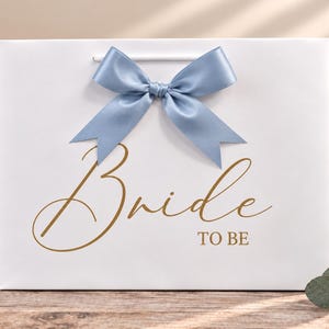 May include: A white rectangular gift bag featuring a dusty blue satin ribbon bow at the top center. The front of the bag displays the text "Bride TO BE" in a gold-colored script and block font. The bag has white cord handles.