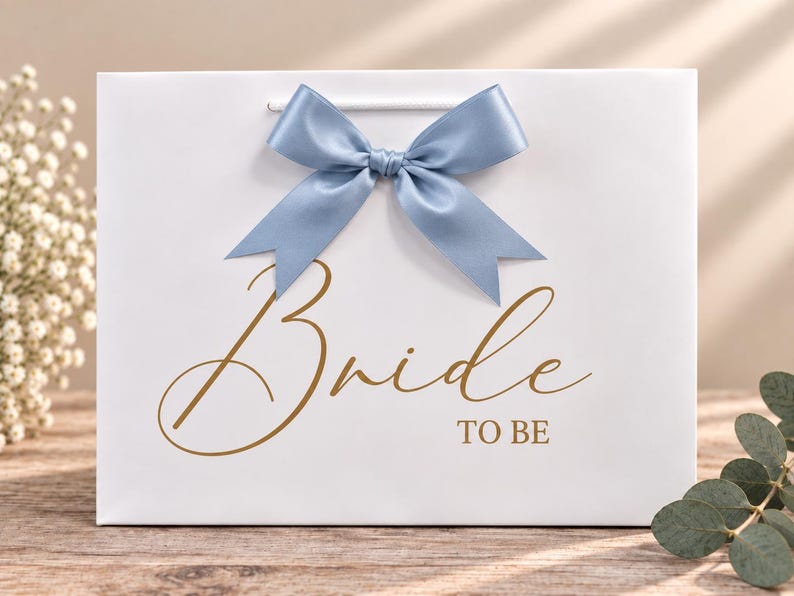 May include: A white rectangular gift bag featuring a dusty blue satin ribbon bow at the top center. The front of the bag displays the text "Bride TO BE" in a gold-colored script and block font. The bag has white cord handles.