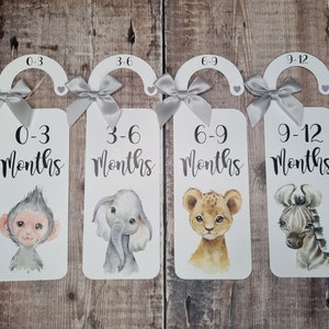 May include: Four white wooden hangers with grey ribbon bows and animal illustrations. Each hanger has a number and text indicating a baby's age in months: 0-3 months, 3-6 months, 6-9 months, and 9-12 months.