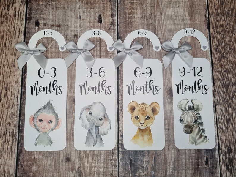 May include: Four white wooden hangers with grey ribbon bows and animal illustrations. Each hanger has a number and text indicating a baby's age in months: 0-3 months, 3-6 months, 6-9 months, and 9-12 months.