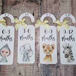 May include: Four white wooden hangers with yellow ribbon bows and animal illustrations. Each hanger has a number and text indicating age ranges for babies: 0-3 months, 3-6 months, 6-9 months, and 9-12 months.