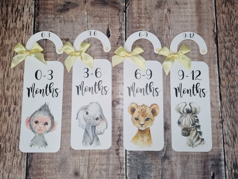 May include: Four white wooden hangers with yellow ribbon bows and animal illustrations. Each hanger has a number and text indicating age ranges for babies: 0-3 months, 3-6 months, 6-9 months, and 9-12 months.
