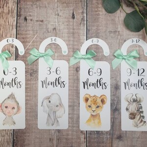 May include: Four white door hangers with green ribbon bows and animal illustrations. Each hanger has a different age range printed on it: 0-3 months, 3-6 months, 6-9 months, and 9-12 months.