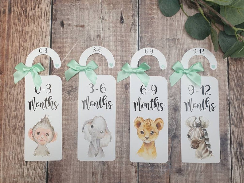 May include: Four white door hangers with green ribbon bows and animal illustrations. Each hanger has a different age range printed on it: 0-3 months, 3-6 months, 6-9 months, and 9-12 months.