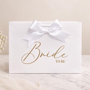 May include: A white rectangular gift bag featuring a large white satin ribbon bow at the top center. The front of the bag displays the text "Bride TO BE" printed in a metallic gold script and block font style across the center.