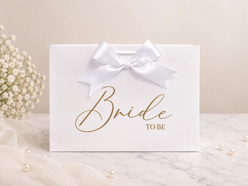 May include: A white rectangular gift bag featuring a large white satin ribbon bow at the top center. The front of the bag displays the text "Bride TO BE" printed in a metallic gold script and block font style across the center.