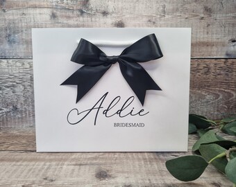 May include: White gift bag with a black satin bow. The bag features the name "Addie" and the word "Bridesmaid" printed in black script. The bag has a white handle.