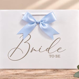 May include: A white gift bag featuring a light blue satin ribbon bow at the top center. The front of the bag displays the text "Bride TO BE" in a dark gray script and block font. The bag has white cord handles and a smooth finish.