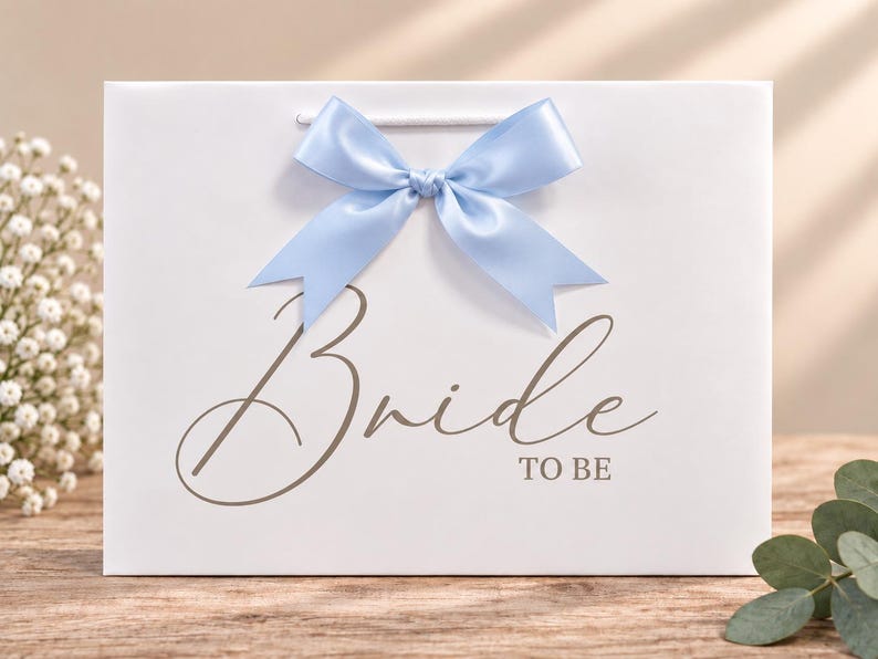 May include: A white gift bag featuring a light blue satin ribbon bow at the top center. The front of the bag displays the text "Bride TO BE" in a dark gray script and block font. The bag has white cord handles and a smooth finish.