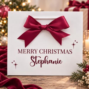 May include: A white gift bag featuring a large, dark red satin ribbon bow at the top. The bag is printed with the dark red text MERRY CHRISTMAS and the name Stephanie in a script font, accented by small four-pointed star graphics on the sides.