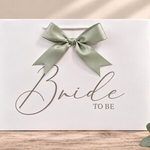 May include: A white rectangular gift bag featuring the text "Bride TO BE" in a dark gray script and block font. The bag is adorned with a sage green satin ribbon tied into a bow at the top center, positioned above the printed text.