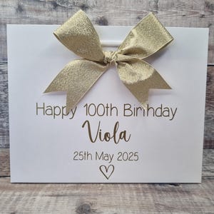 Puede incluir: Una bolsa de regalo blanca con un lazo dorado. La bolsa tiene el texto "Happy 100th Birthday Viola" y la fecha "25 de mayo de 2025" impresos en dorado. Un s&iacute;mbolo de coraz&oacute;n est&aacute; debajo de la fecha.