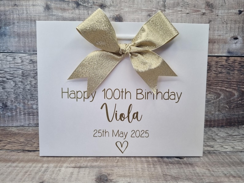 Puede incluir: Una bolsa de regalo blanca con un lazo dorado. La bolsa tiene el texto "Happy 100th Birthday Viola" y la fecha "25 de mayo de 2025" impresos en dorado. Un s&iacute;mbolo de coraz&oacute;n est&aacute; debajo de la fecha.