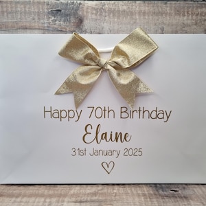 Puede incluir: Bolsa de regalo blanca con un lazo dorado y el texto "Happy 70th Birthday Elaine 31st January 2025" en letras doradas. Un s&iacute;mbolo de coraz&oacute;n est&aacute; debajo de la fecha. La bolsa est&aacute; sobre una superficie de madera.