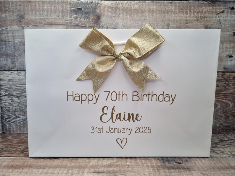 Puede incluir: Bolsa de regalo blanca con un lazo dorado y el texto "Happy 70th Birthday Elaine 31st January 2025" en letras doradas. Un s&iacute;mbolo de coraz&oacute;n est&aacute; debajo de la fecha. La bolsa est&aacute; sobre una superficie de madera.