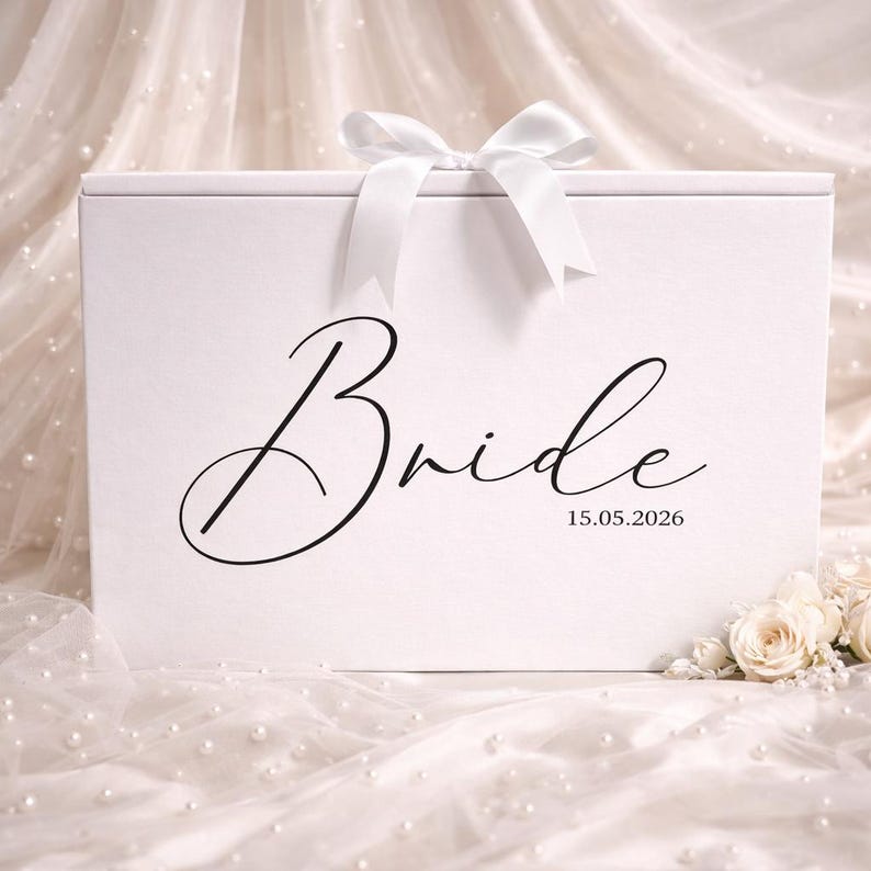 May include: A white gift box with a white satin bow. The word "Bride" is written in elegant script, with the date 15.05.2026 below. The box is on a white fabric with pearls and flowers.