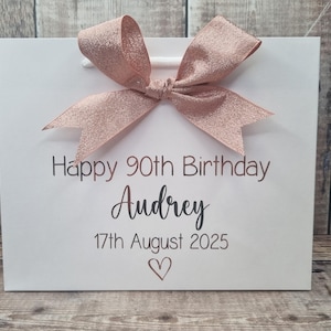 Puede incluir: Bolsa de regalo blanca con un lazo de purpurina oro rosa. La bolsa tiene el texto "Happy 90th Birthday Audrey 17th August 2025" y un s&iacute;mbolo de coraz&oacute;n impreso en color marr&oacute;n oscuro.