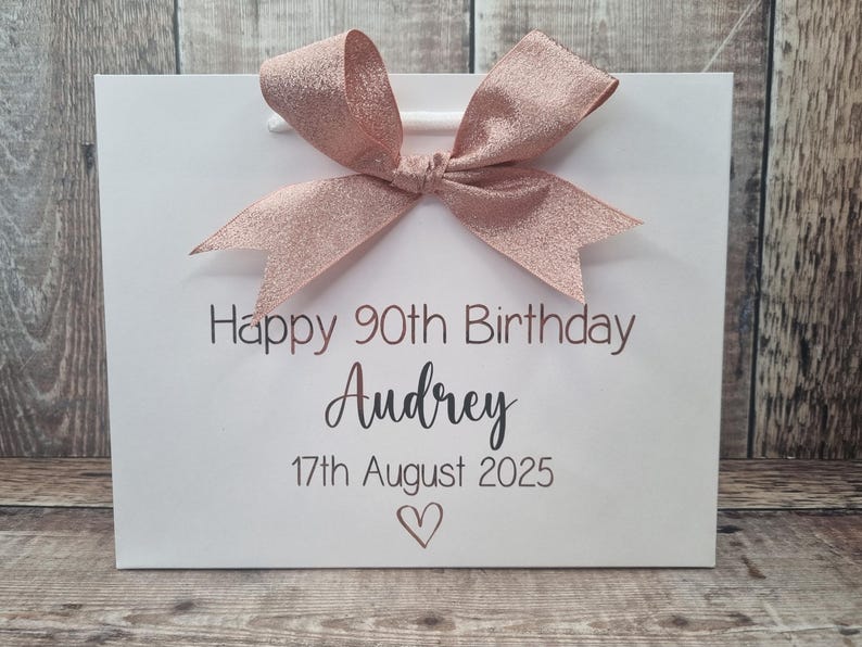 Puede incluir: Bolsa de regalo blanca con un lazo de purpurina oro rosa. La bolsa tiene el texto "Happy 90th Birthday Audrey 17th August 2025" y un s&iacute;mbolo de coraz&oacute;n impreso en color marr&oacute;n oscuro.