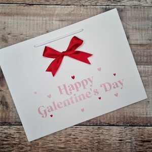 May include: White gift bag with a red bow and the text "Happy Galentine's Day" in pink letters. The bag is decorated with small pink hearts.