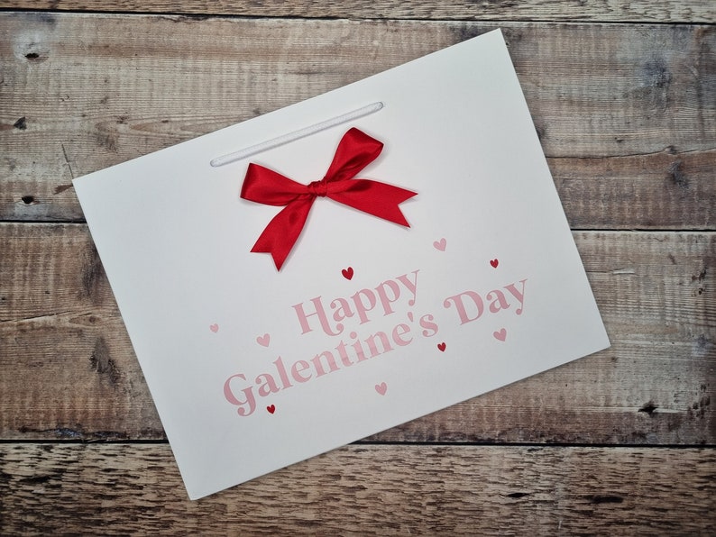May include: White gift bag with a red bow and the text "Happy Galentine's Day" in pink letters. The bag is decorated with small pink hearts.