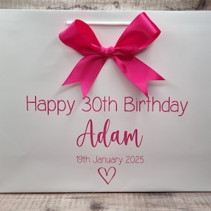 Puede incluir: Bolsa de regalo blanca con un lazo rosa brillante y texto a juego. El texto dice "Feliz 30 cumplea&ntilde;os Adam 19 de enero de 2025" con un s&iacute;mbolo de coraz&oacute;n. La bolsa tiene un asa de cord&oacute;n blanco.