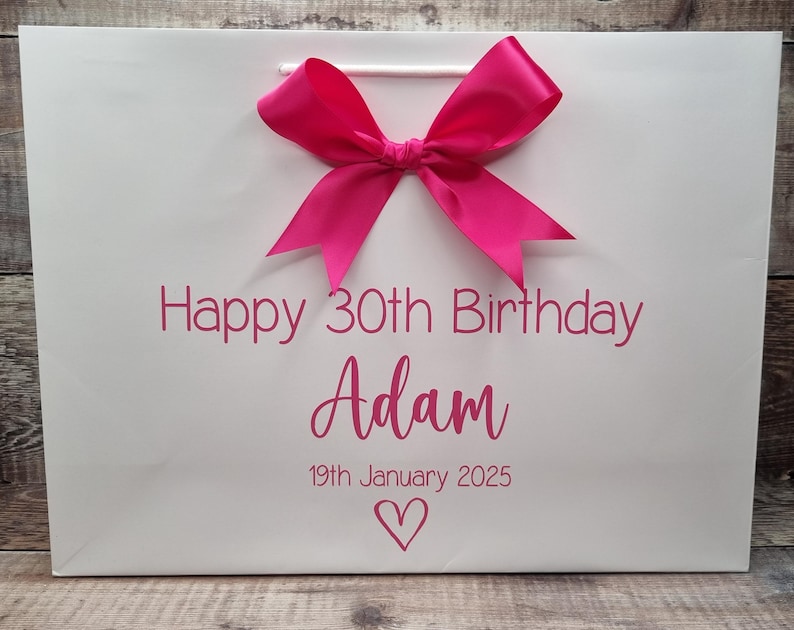 Puede incluir: Bolsa de regalo blanca con un lazo rosa brillante y texto a juego. El texto dice "Feliz 30 cumplea&ntilde;os Adam 19 de enero de 2025" con un s&iacute;mbolo de coraz&oacute;n. La bolsa tiene un asa de cord&oacute;n blanco.