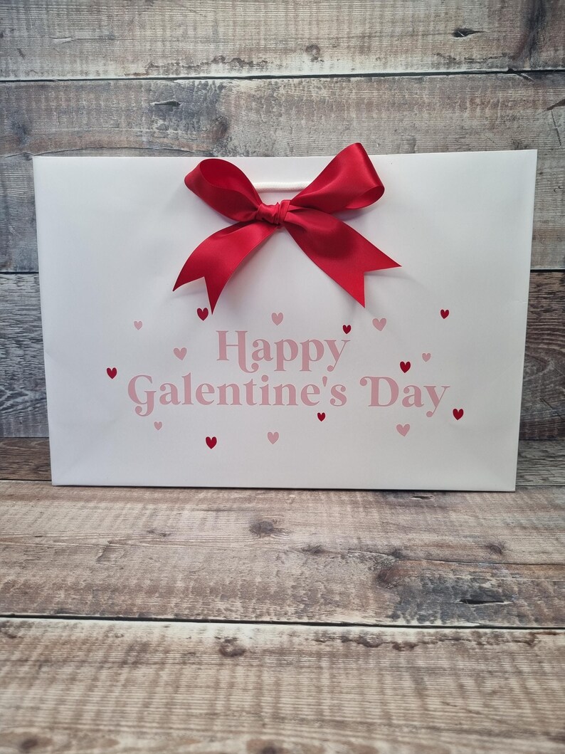 May include: White gift bag with a red bow and the text "Happy Galentine's Day" in pink lettering. The bag is decorated with small pink hearts.