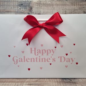 May include: White gift bag with a red bow and the text "Happy Galentine's Day" in pink letters with pink hearts scattered around the text.
