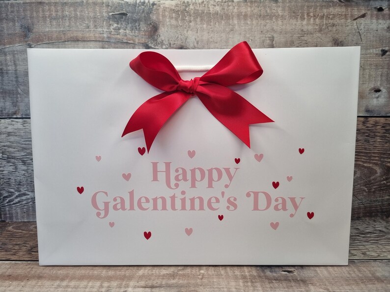 May include: White gift bag with a red bow and the text "Happy Galentine's Day" in pink letters with pink hearts scattered around the text.