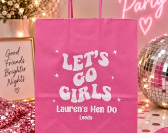 Puede incluir: Una bolsa de regalo de papel rosa brillante con dos asas a juego. La parte frontal presenta texto blanco que dice Let's Go Girls, Lauren's Hen Do y Leeds, acentuado con pequeños iconos de estrellas blancas impresos en el centro.
