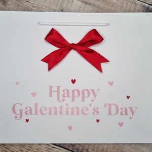 May include: White gift bag with a red bow and the text "Happy Galentine's Day" in pink lettering. The bag is decorated with small pink hearts.