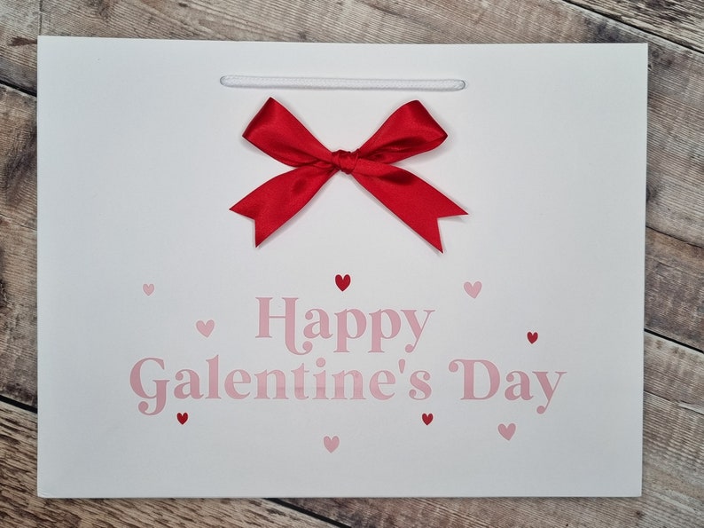May include: White gift bag with a red bow and the text "Happy Galentine's Day" in pink lettering. The bag is decorated with small pink hearts.