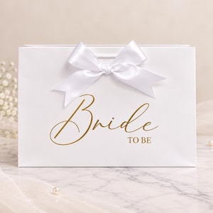 May include: A rectangular white gift bag featuring a large white satin ribbon bow at the top center. The front of the bag displays the text "Bride TO BE" printed in a metallic gold cursive and block font style.