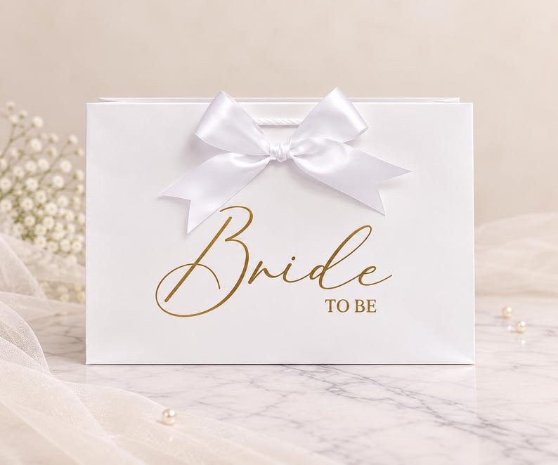 May include: A rectangular white gift bag featuring a large white satin ribbon bow at the top center. The front of the bag displays the text "Bride TO BE" printed in a metallic gold cursive and block font style.