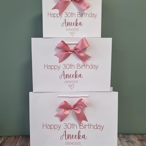 Puede incluir: Tres bolsas de regalo blancas apiladas, cada una adornada con un lazo de sat&eacute;n rosa. Las bolsas presentan el texto "Happy 30th Birthday Aneeka 01/04/2025" con un coraz&oacute;n, ideal para una celebraci&oacute;n.