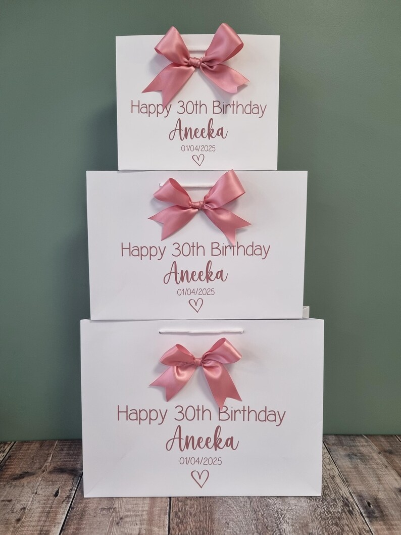Puede incluir: Tres bolsas de regalo blancas apiladas, cada una adornada con un lazo de sat&eacute;n rosa. Las bolsas presentan el texto "Happy 30th Birthday Aneeka 01/04/2025" con un coraz&oacute;n, ideal para una celebraci&oacute;n.
