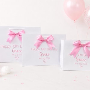 Puede incluir: Tres bolsas de regalo blancas con lazos rosas, personalizadas con "Happy 18th Birthday Grace" y la fecha. Una bola de panal rosa, globos y confeti completan la escena festiva. Las bolsas son de diferentes tama&ntilde;os.