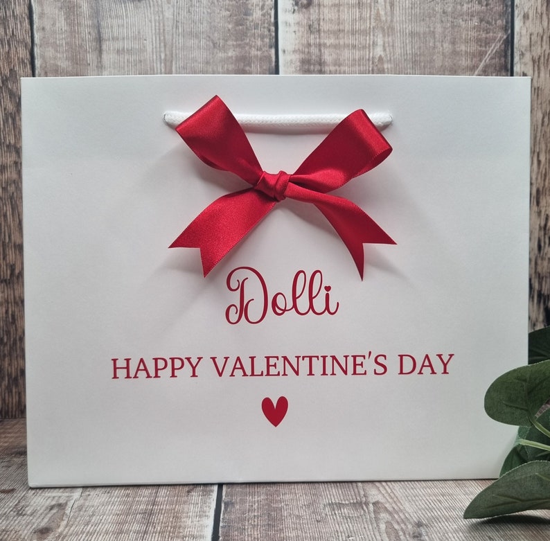 May include: White gift bag with a red bow and the text "Dolli Happy Valentine's Day" with a red heart.
