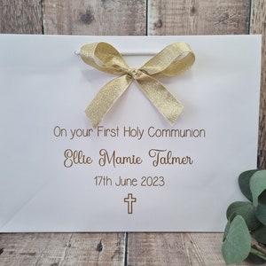 Op de afbeelding: Een witte cadeauzak met een gouden glitterstrik aan de bovenkant. De tekst luidt On your First Holy Communion, Ellie Mamie Talmer, 17th June 2023, met een klein gouden kruisicoon onder de datum gedrukt in een eenvoudig lettertype.