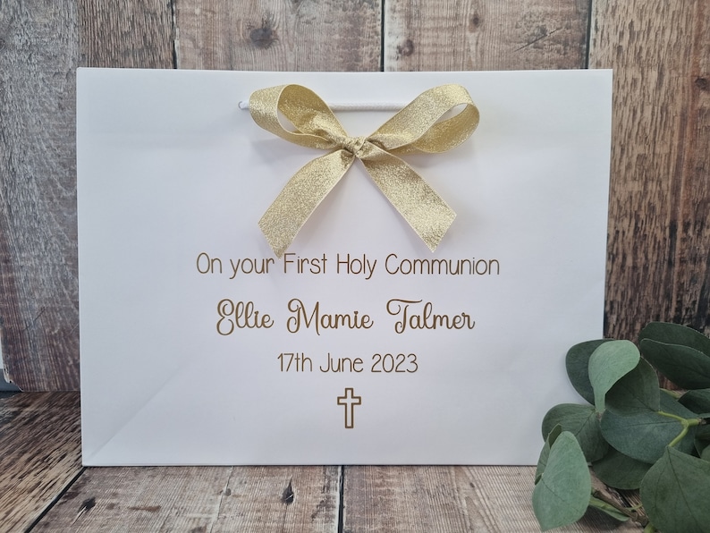 Op de afbeelding: Een witte cadeauzak met een gouden glitterstrik aan de bovenkant. De tekst luidt On your First Holy Communion, Ellie Mamie Talmer, 17th June 2023, met een klein gouden kruisicoon onder de datum gedrukt in een eenvoudig lettertype.