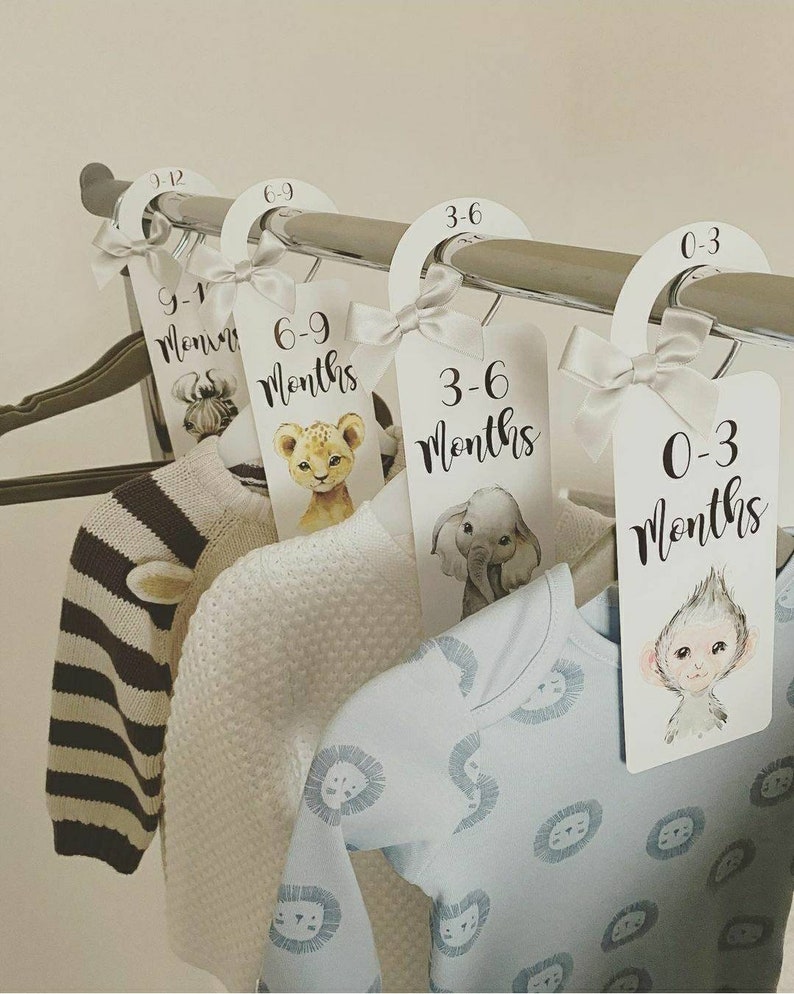 May include: Four white hangers with animal illustrations and text indicating age ranges for baby clothes. The hangers are labelled 0-3 months, 3-6 months, 6-9 months, and 9-12 months. The hangers are hanging on a silver clothes rack.