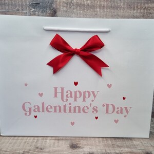 May include: White gift bag with a red bow and the text "Happy Galentine's Day" in pink lettering. The bag is decorated with small pink hearts.