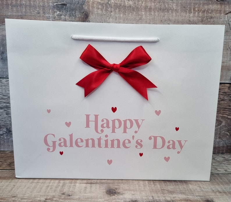 May include: White gift bag with a red bow and the text "Happy Galentine's Day" in pink lettering. The bag is decorated with small pink hearts.