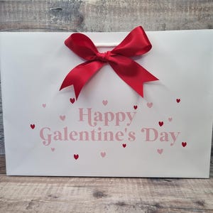 May include: White gift bag with a red bow and the text "Happy Galentine's Day" in pink letters. The bag is decorated with small pink and red hearts.