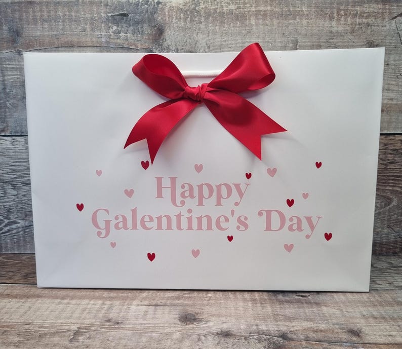 May include: White gift bag with a red bow and the text "Happy Galentine's Day" in pink letters. The bag is decorated with small pink and red hearts.